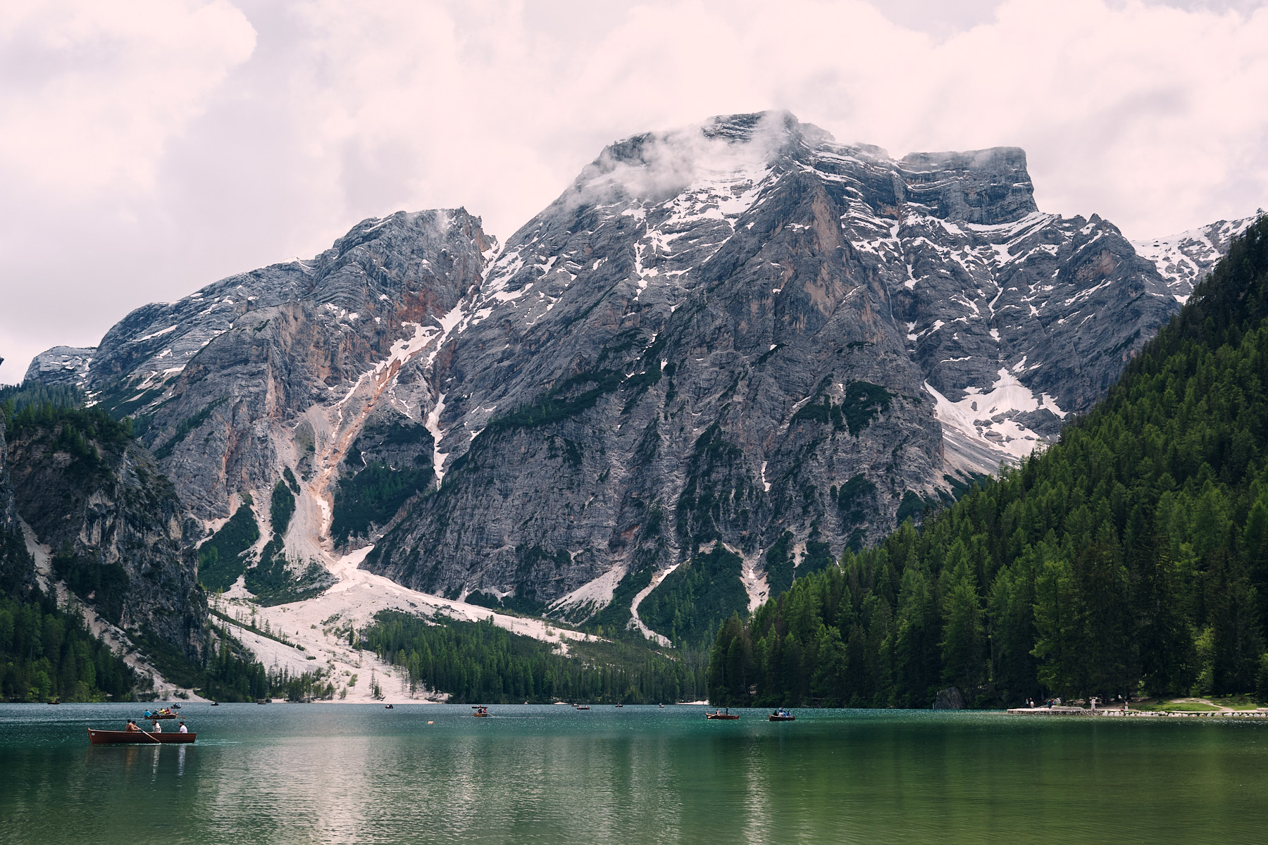Steep limestone mountains with snow patches rise above a turquoise alpine lake where small boats drift near forested shores.