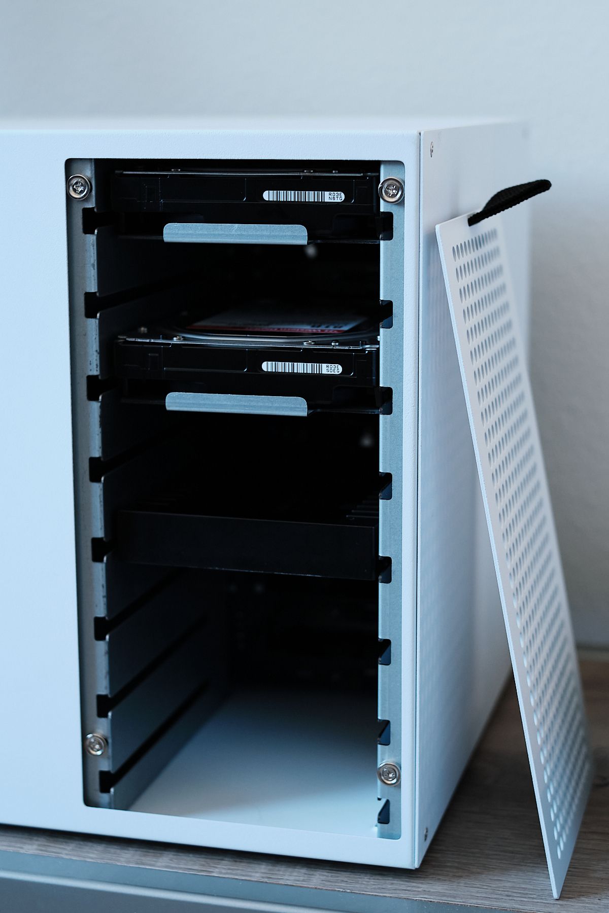 The NAS case with its perforated front panel removed and leaning against the side, revealing two hard drives with silver mounting tabs and the SSD adapter bracket installed in the drive bays, with the lower bays left empty.