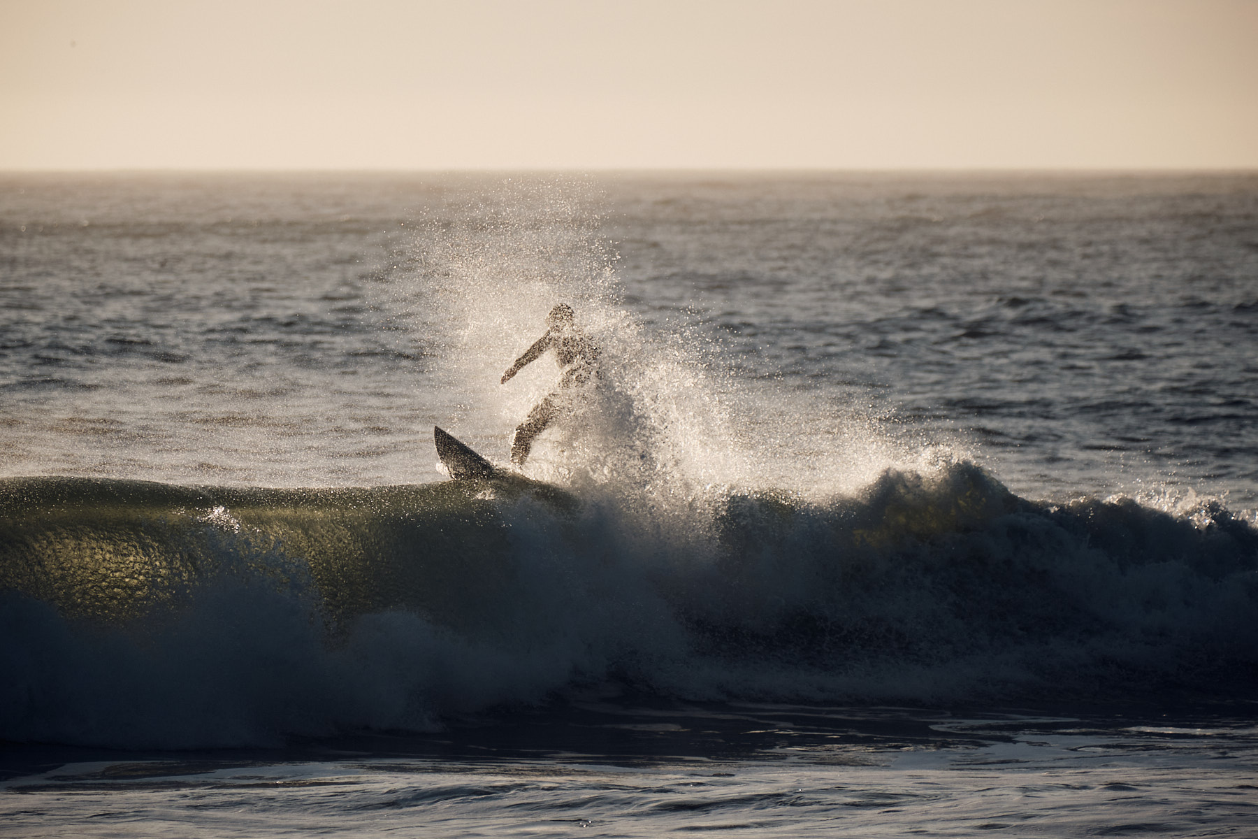 A surfer performs an aerial maneuver above a breaking wave during sunset, creating a dramatic spray of water against a dusky ocean backdrop.
