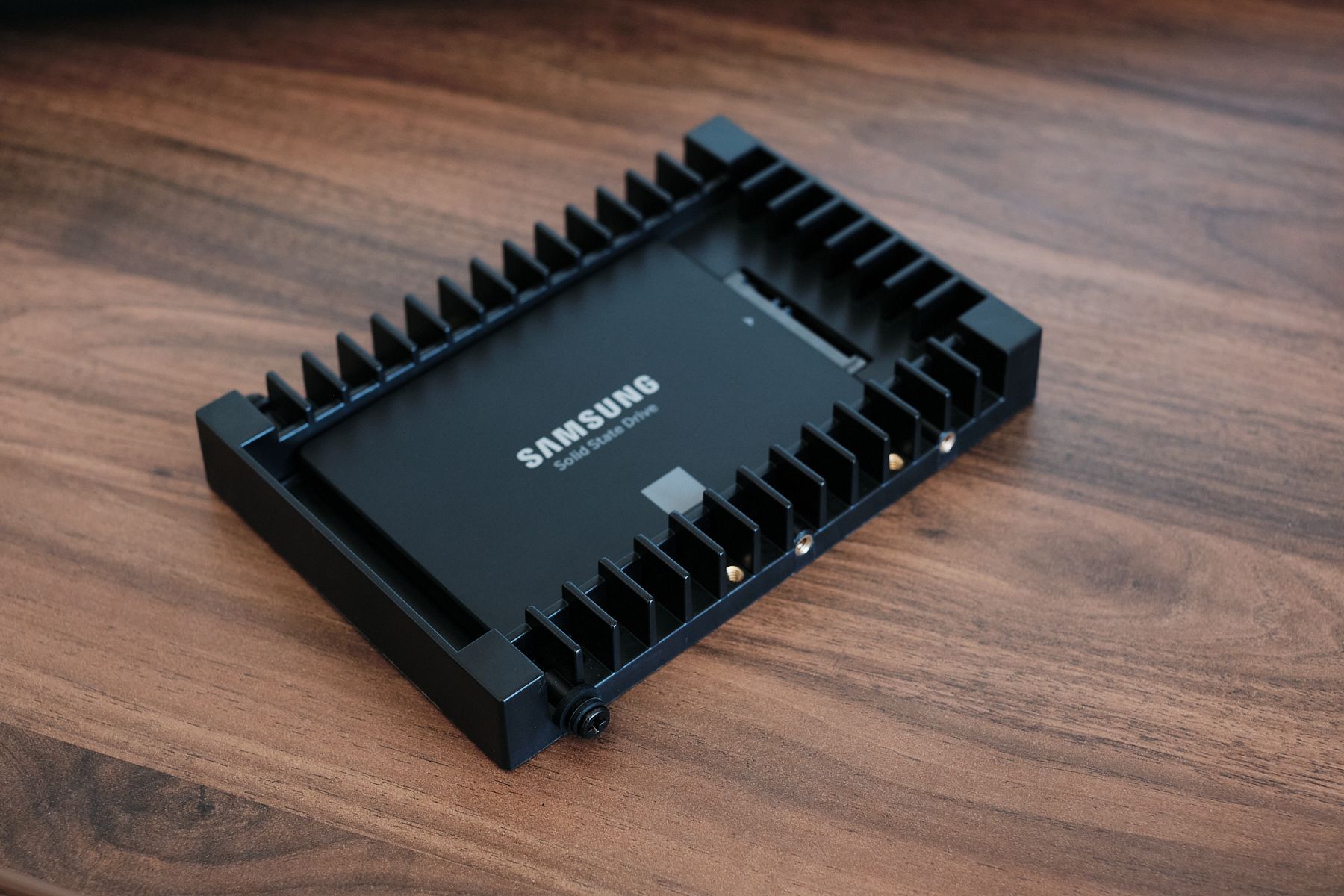 A Samsung 2.5-inch SSD seated in a black 3.5-inch adapter bracket with a rubber-grommeted mounting screw on its side, resting on a wooden desk.